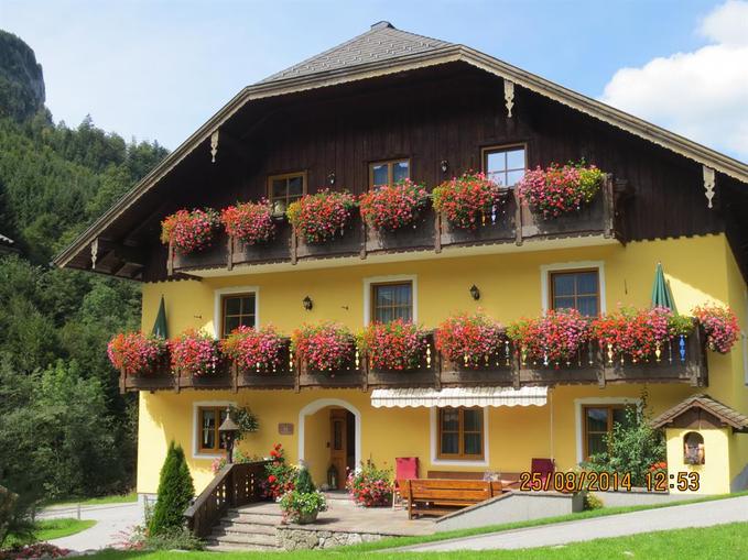 A charming yellow house with many flower boxes on the balconies. The surroundings are green and natural, with trees and a clear sky in the background.
