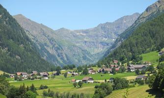 A picturesque landscape with a small village in a green valley. Tall mountains surround the scene under a clear blue sky.