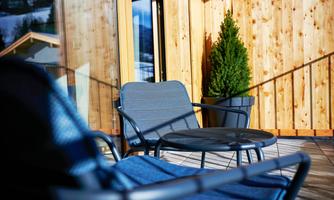 A cozy balcony with two chairs and a small table. In the background, a green plant and wooden paneling are visible.