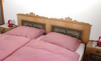 A bright bedroom with two single beds and pink bedding. Intricately designed headboards hang on the wall, next to which are red bedside lamps.