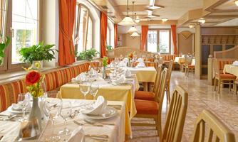 An elegant restaurant with tablecloths in yellow and orange. The rooms are bright and friendly with plants and large windows.