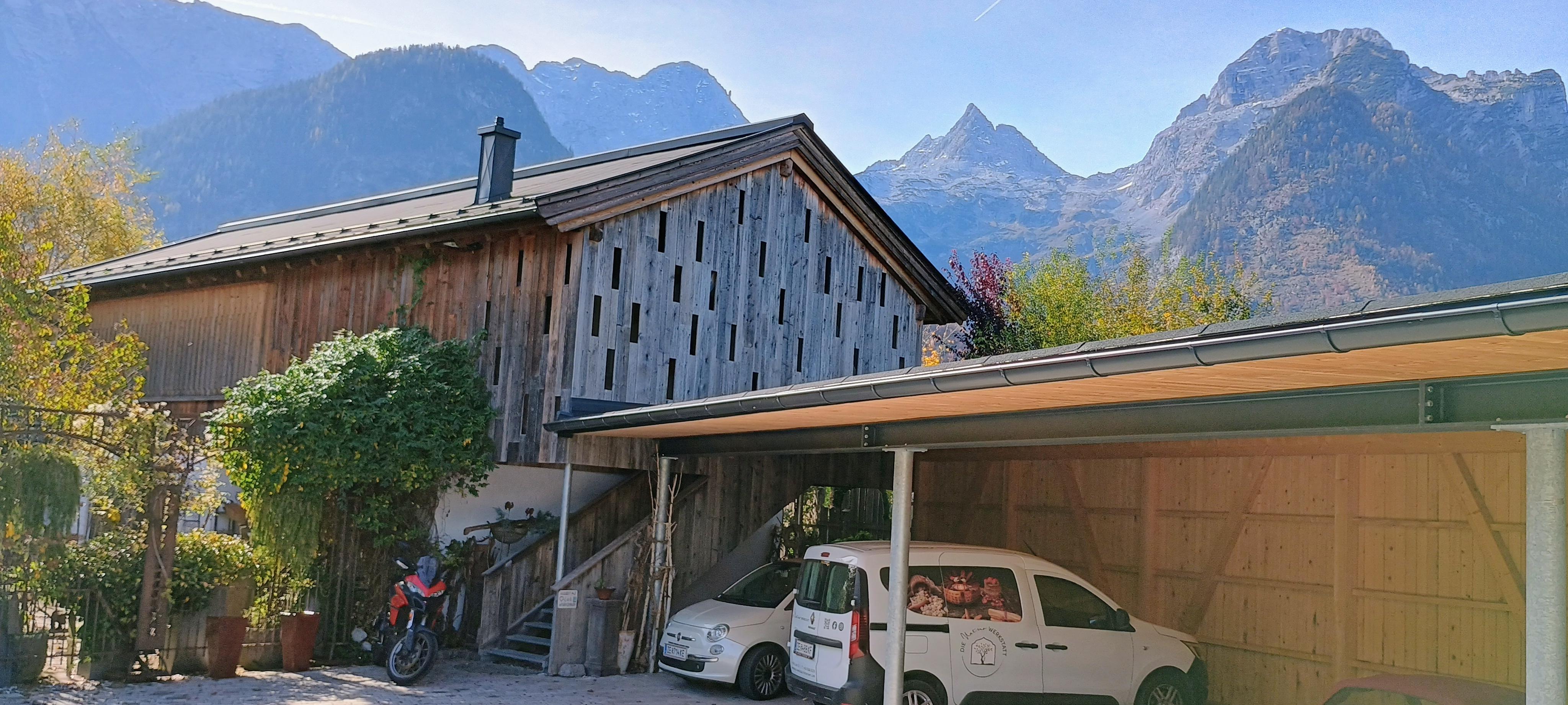 A rustic house with wooden siding, surrounded by mountains. In the foreground, two vehicles are parked next to the garage.