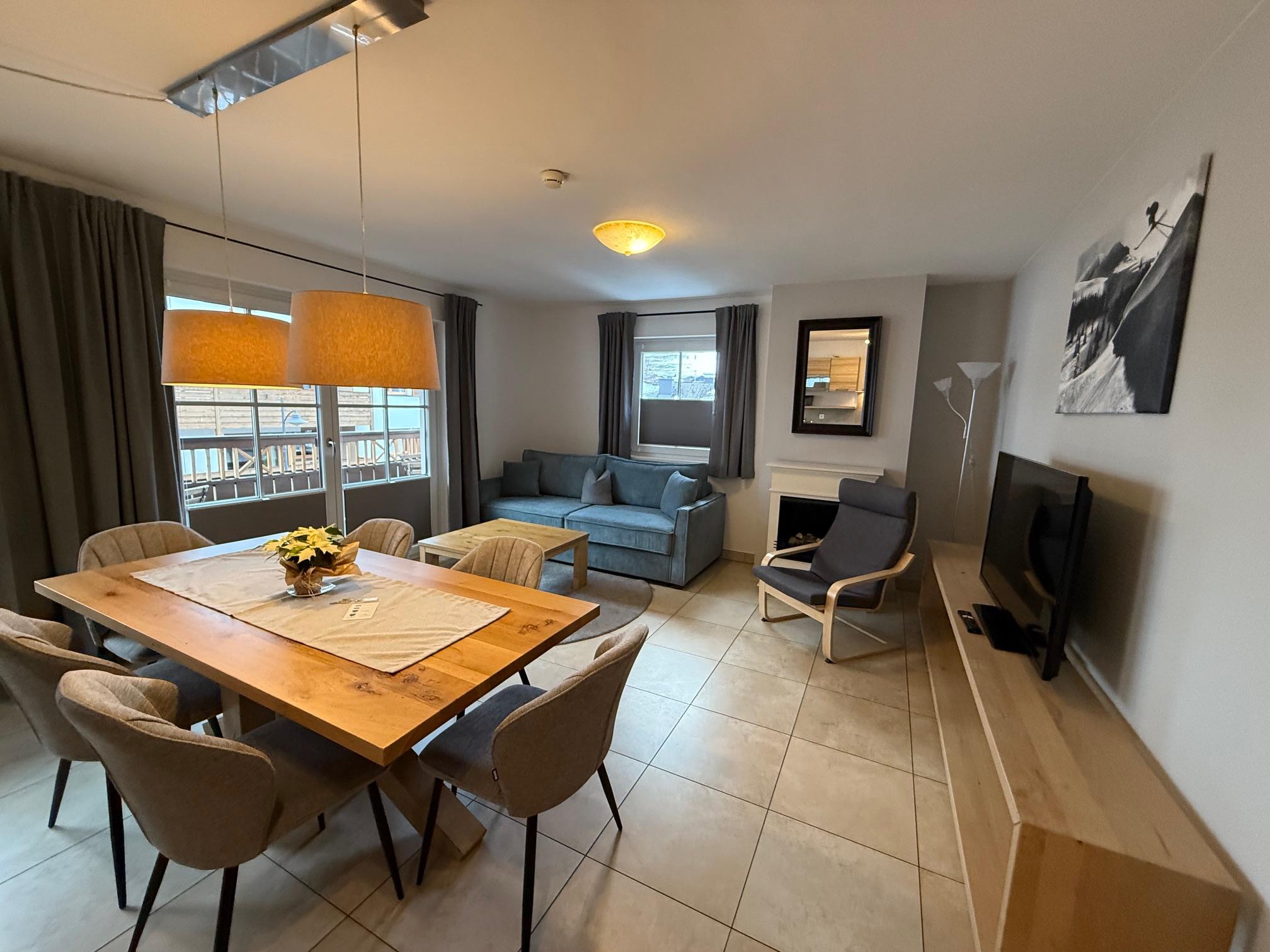 A modern living room with a dining table and six chairs. A cozy sofa and a television are also visible.