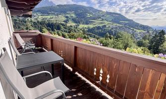 A balcony with two chairs and a table offers a stunning view of the mountains and the valley. The landscape is green and characterized by gentle hills.