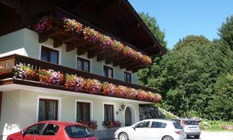 A cozy house with a balcony full of colorful flowers. In the foreground, several cars are parked in a parking lot.