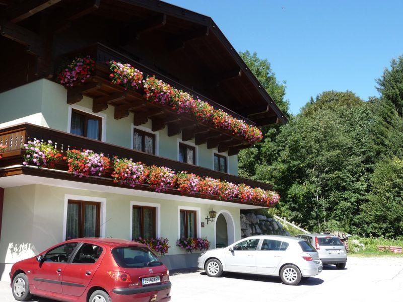 A cozy house with a balcony full of colorful flowers. In the foreground, several cars are parked in a parking lot.