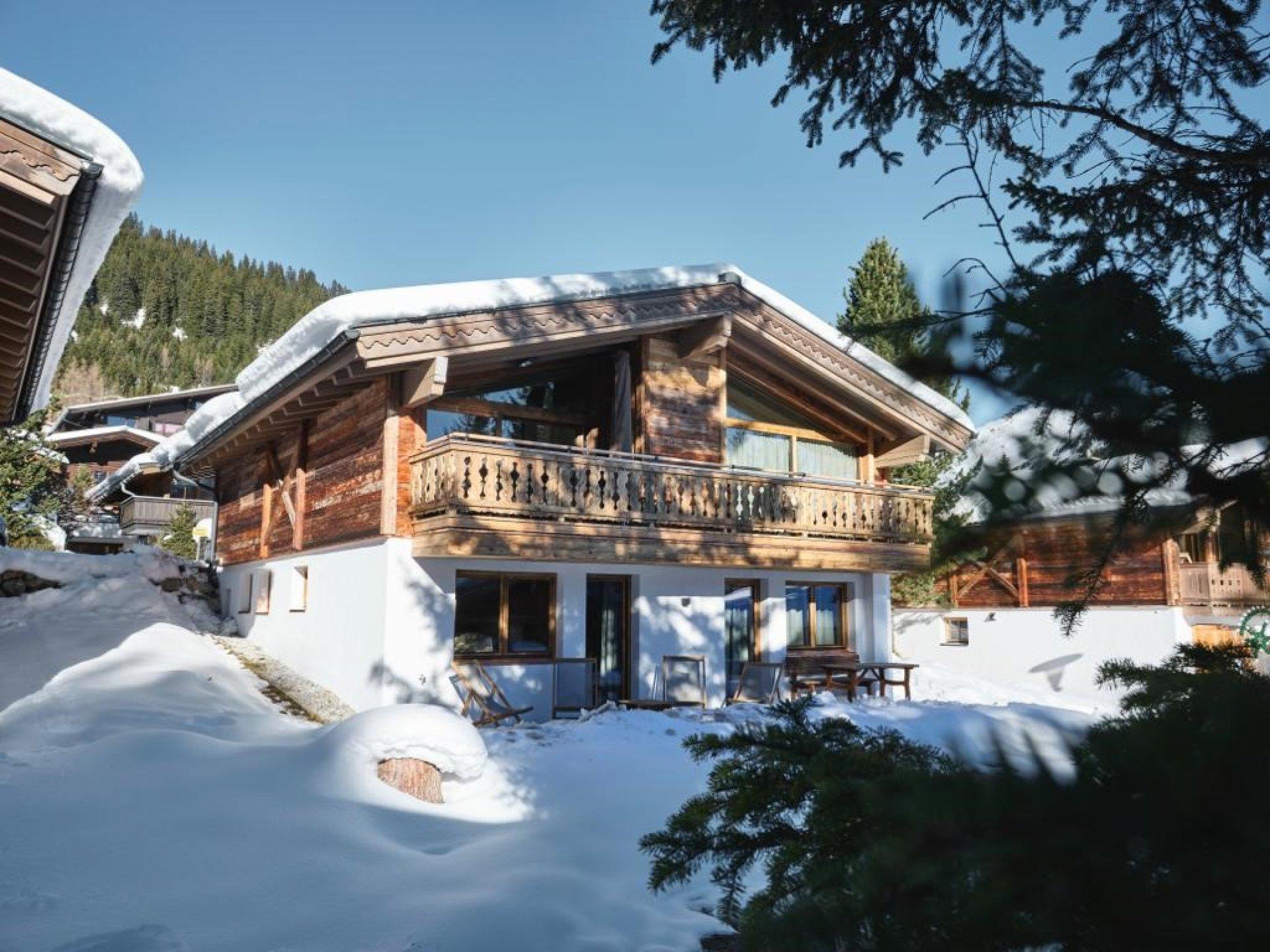 A charming wooden house in the snow, surrounded by snow-covered mountains. The sunlight makes the scene appear bright and inviting.
