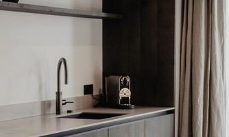 Modern kitchen unit with dark wood surfaces and an elegant faucet. A window with a curtain provides natural light in the room.