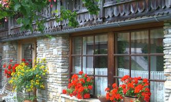 A charming house with stone walls and wooden cladding. Bright red geraniums and yellow flowers bloom above the windows.