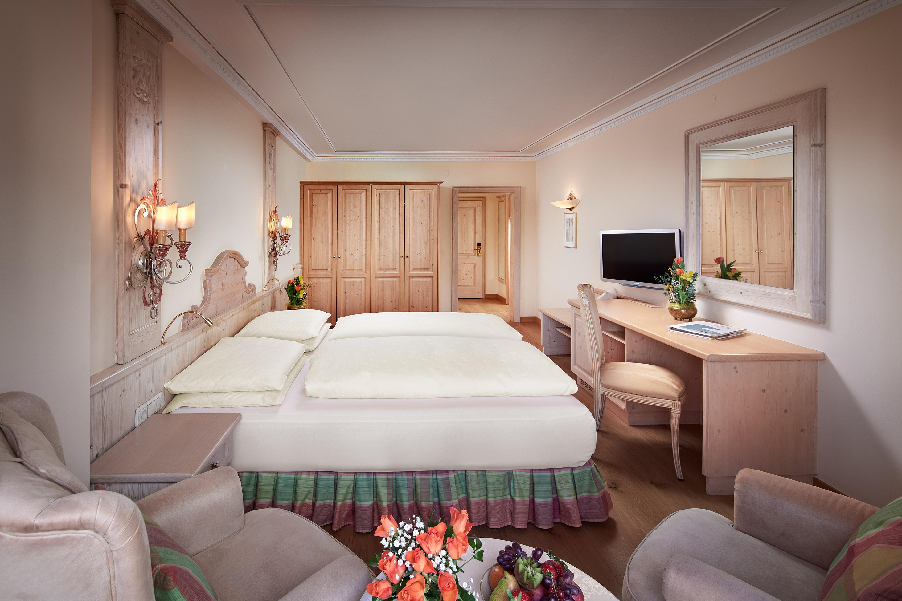An elegant hotel room with a large double bed and bright decor. The room includes a desk, a television, and a cozy seating area.