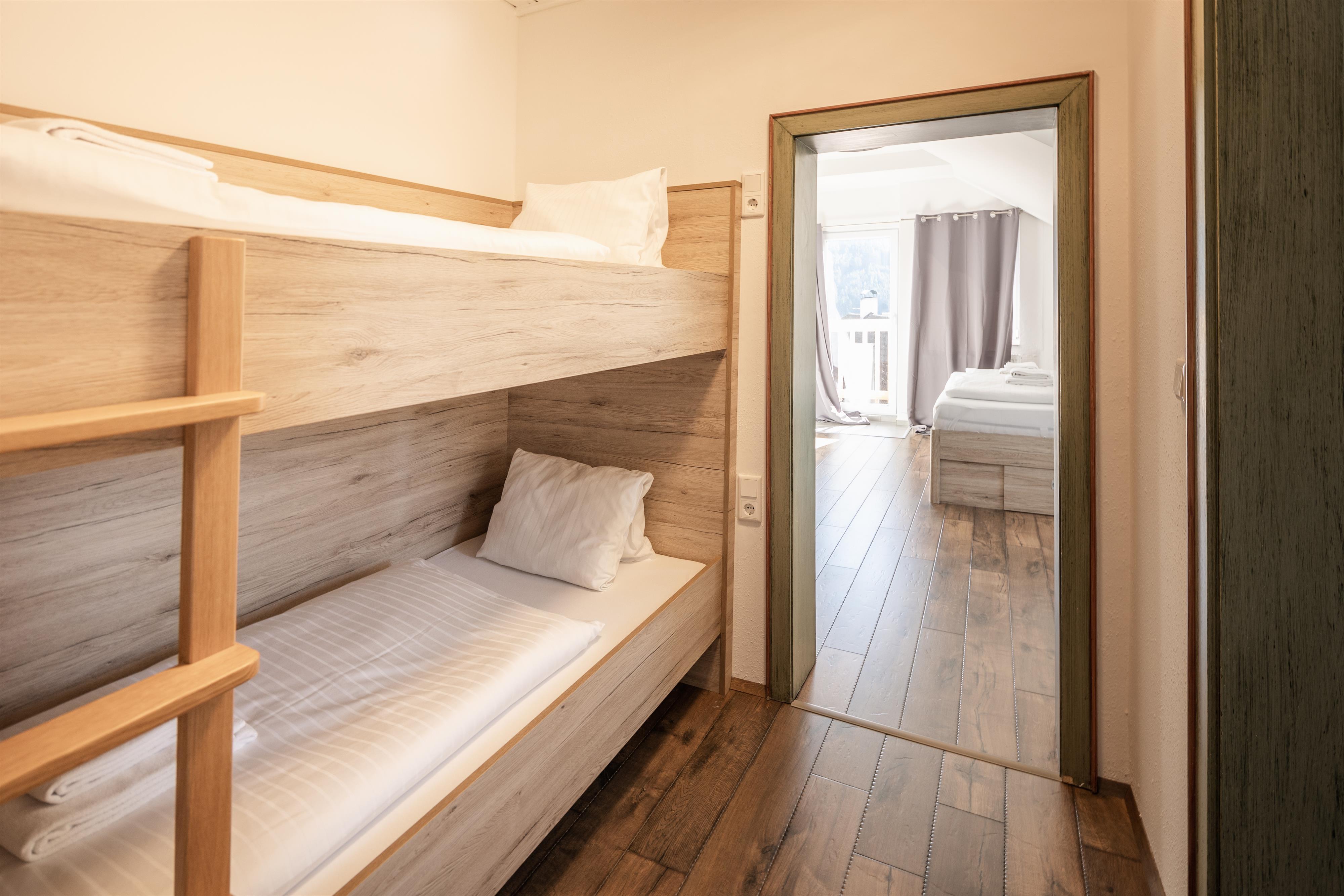 A simple bedroom with a wooden bunk bed. In the background, another room with two beds is visible.
