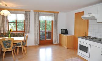 A bright and inviting living room with a kitchen corner. There is a dining area with wooden chairs and a view outside through large windows.