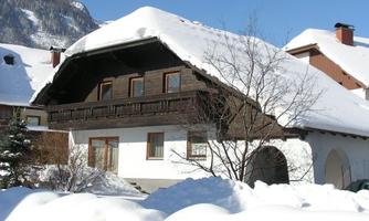 A traditional, charming house with a snow-covered roof. The surroundings are wintry with plenty of snow and a clear blue sky.