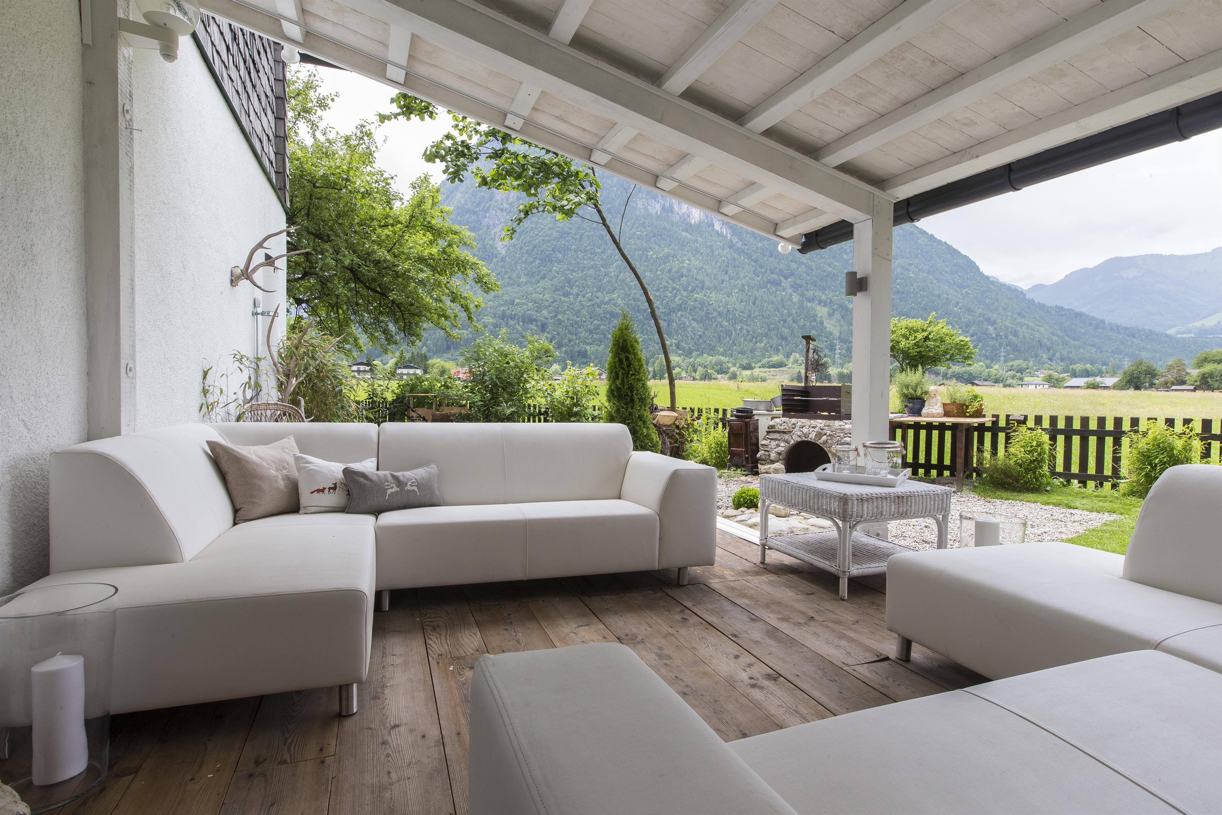 A modern terrace with a white seating area and a table. In the background, green hills and trees can be seen.