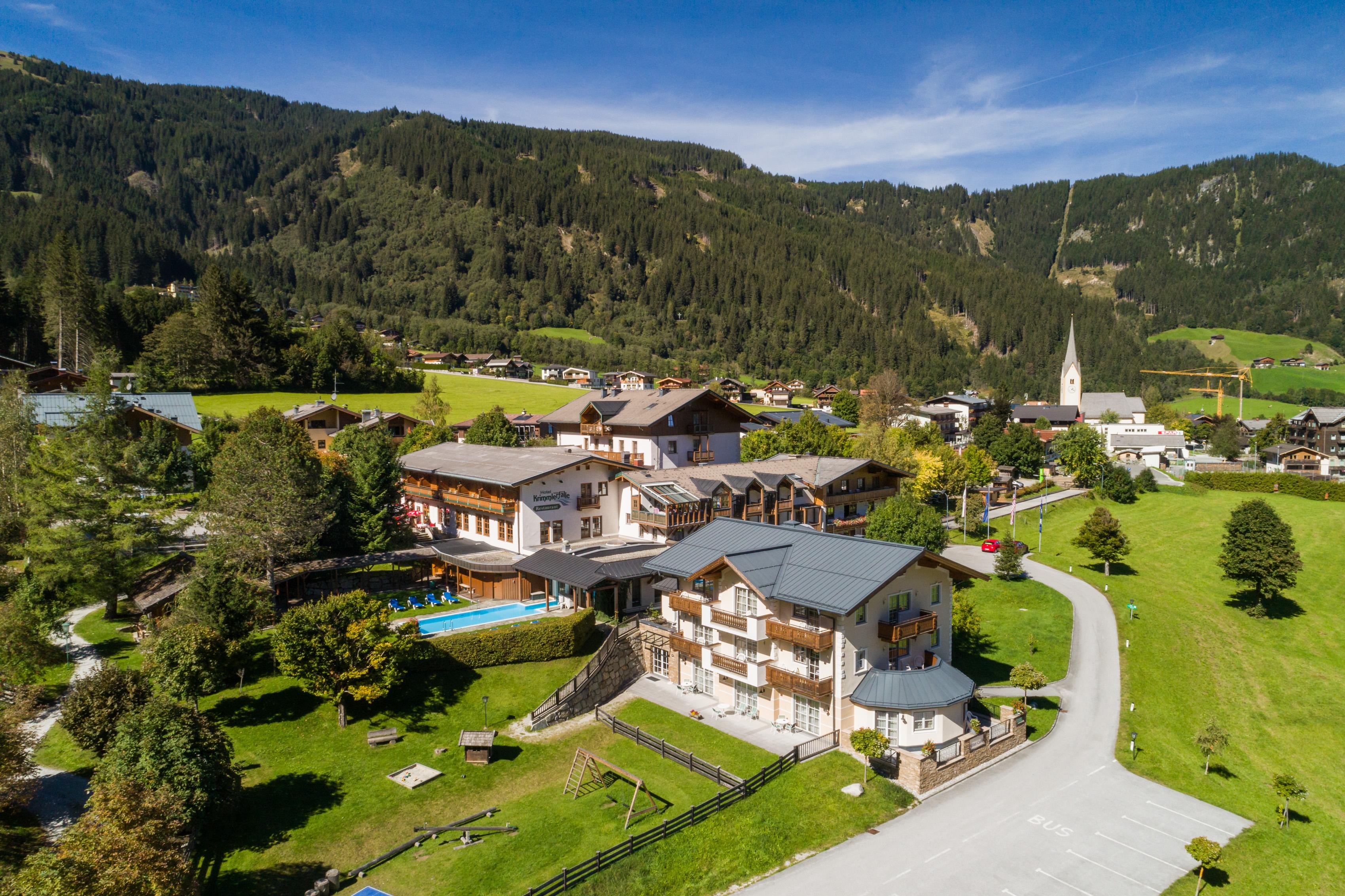A picturesque Alpine landscape with various buildings and green meadows. In the background, mountains and a clear blue sky can be seen.