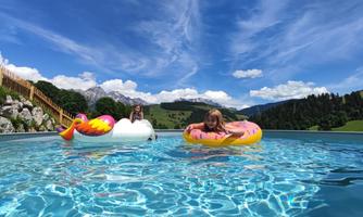 Two girls are swimming in a pool with colorful floaties. In the background, there are green hills and a blue sky with some clouds.