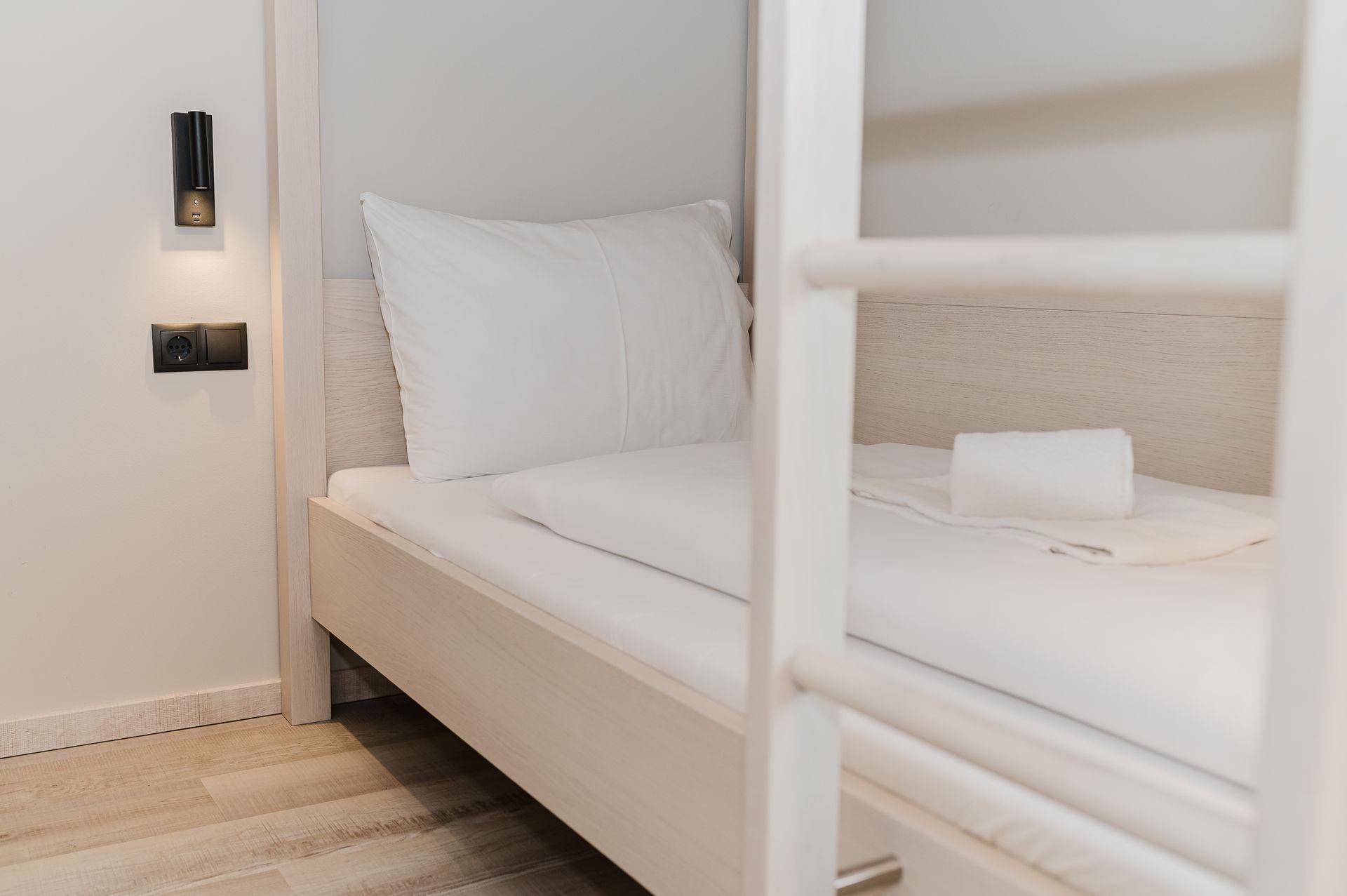 A simple, clean room with a single bed and a white pillow. On the wall, there is a reading lamp.
