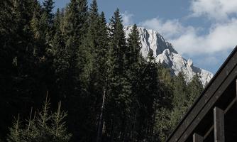 A wooden building at the foot of a wooded hill, with snow-capped mountains in the background. The sky is clear with a few clouds.