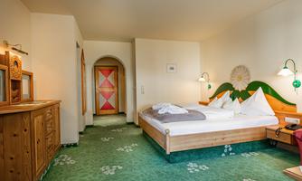 A bright bedroom with a double bed, wooden furniture, and a green carpet. The walls are neutral, and there are decorative lamps as well as a colorful door design.