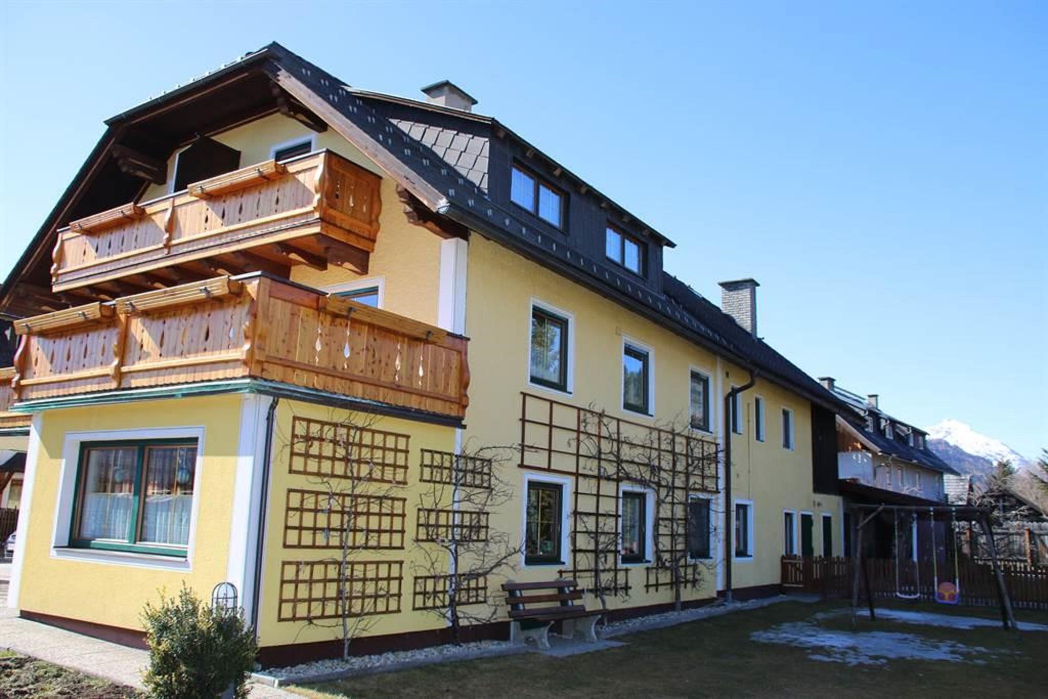 A yellow house with a balcony and wooden decorations. The sky is clear and blue, and the garden is well maintained.