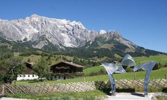 A modern sculpture stands in front of an impressive mountain landscape. In the background, green meadows and traditional houses are visible.