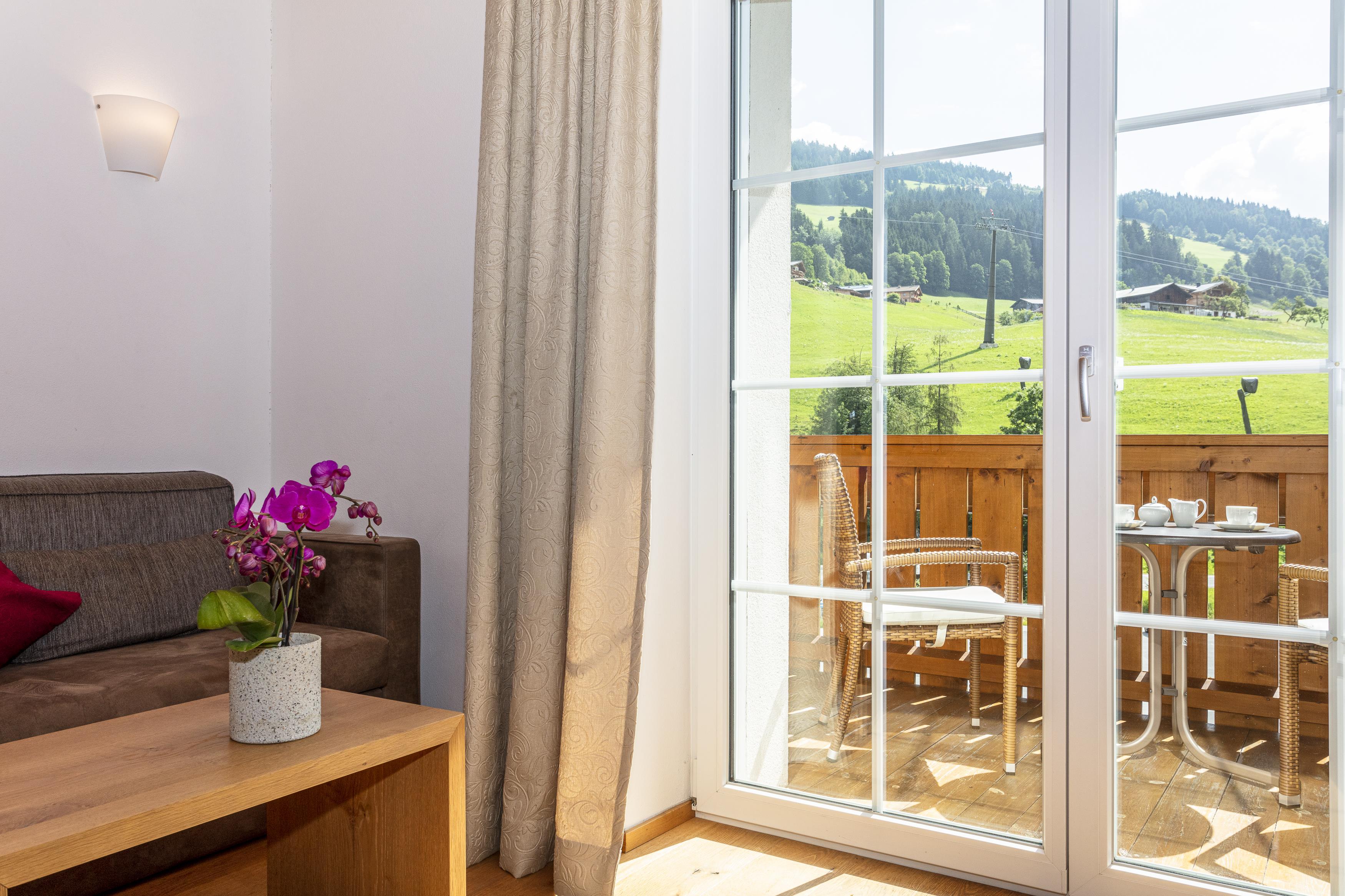 A cozy living room with a view of the green landscape. Outside, there is a small table with chairs on the balcony.