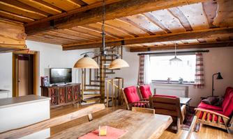A cozy kitchen-living room with wooden ceilings and a large dining table. Red armchairs and a staircase create an inviting atmosphere.