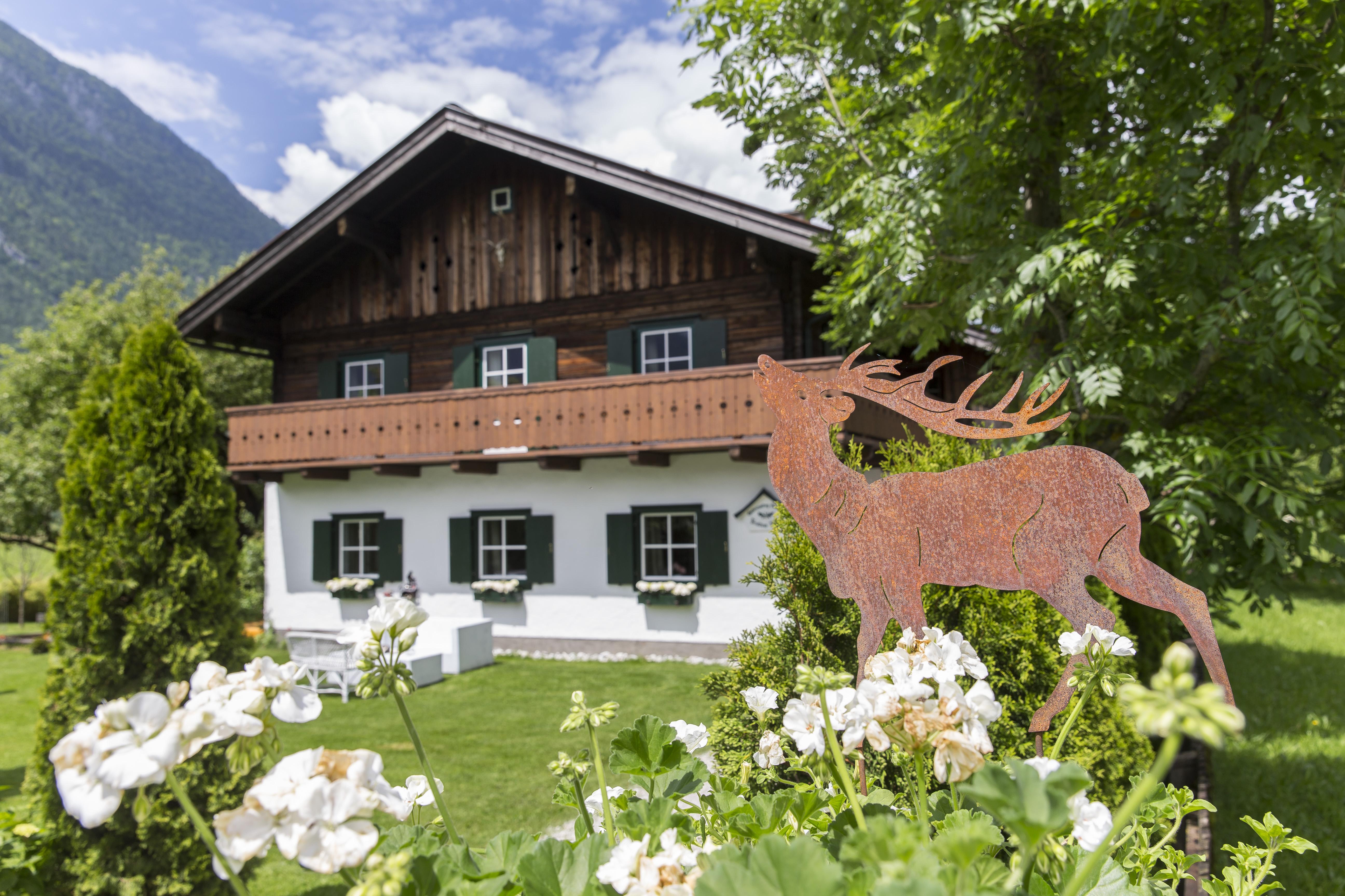 A traditional wooden house surrounded by green trees and flowers. In the foreground stands a decorative metal figure of a deer.