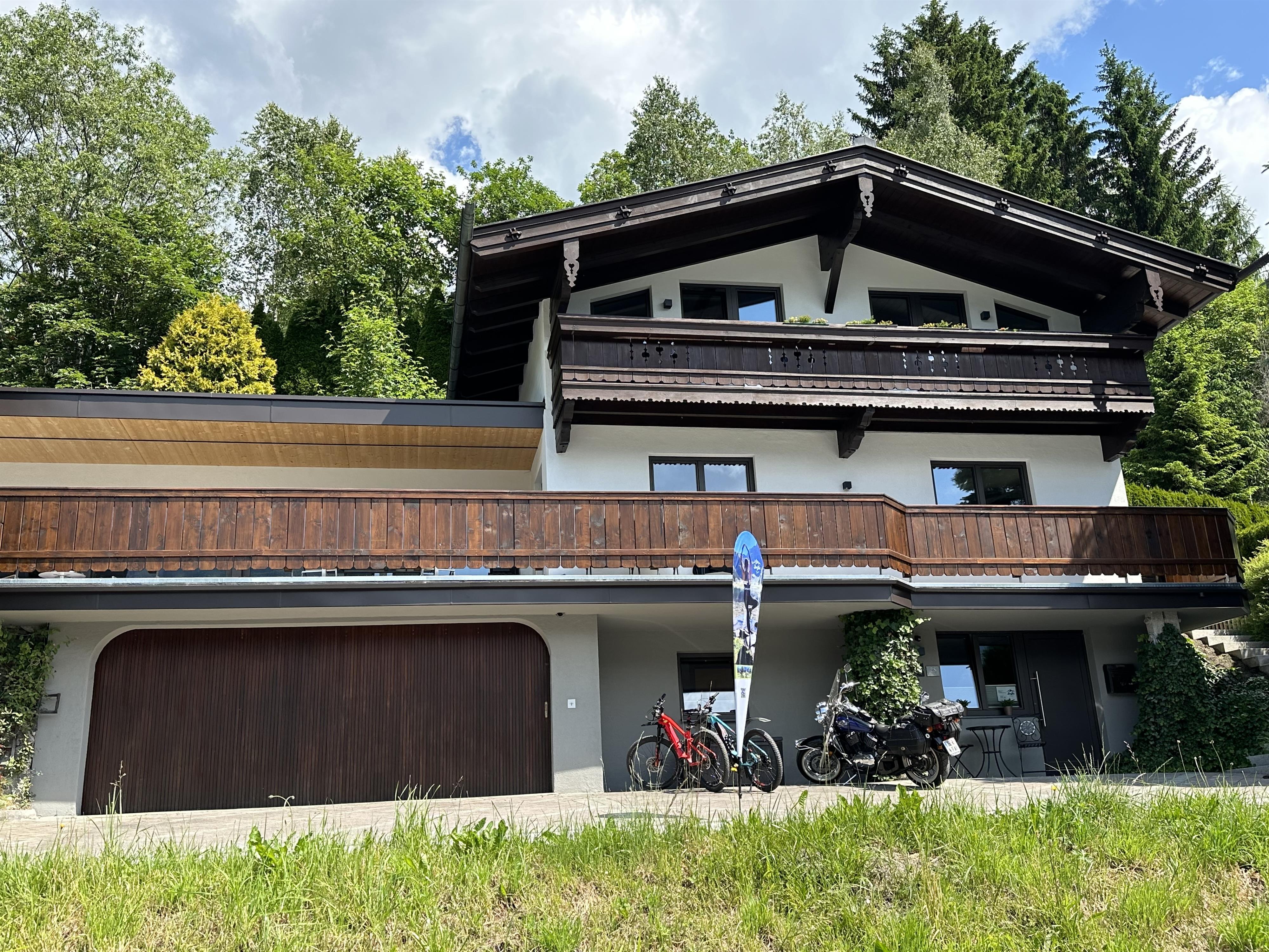 A modern house in alpine style with a large balcony and a garage. In front of the house are two bicycles, surrounded by a green meadow.