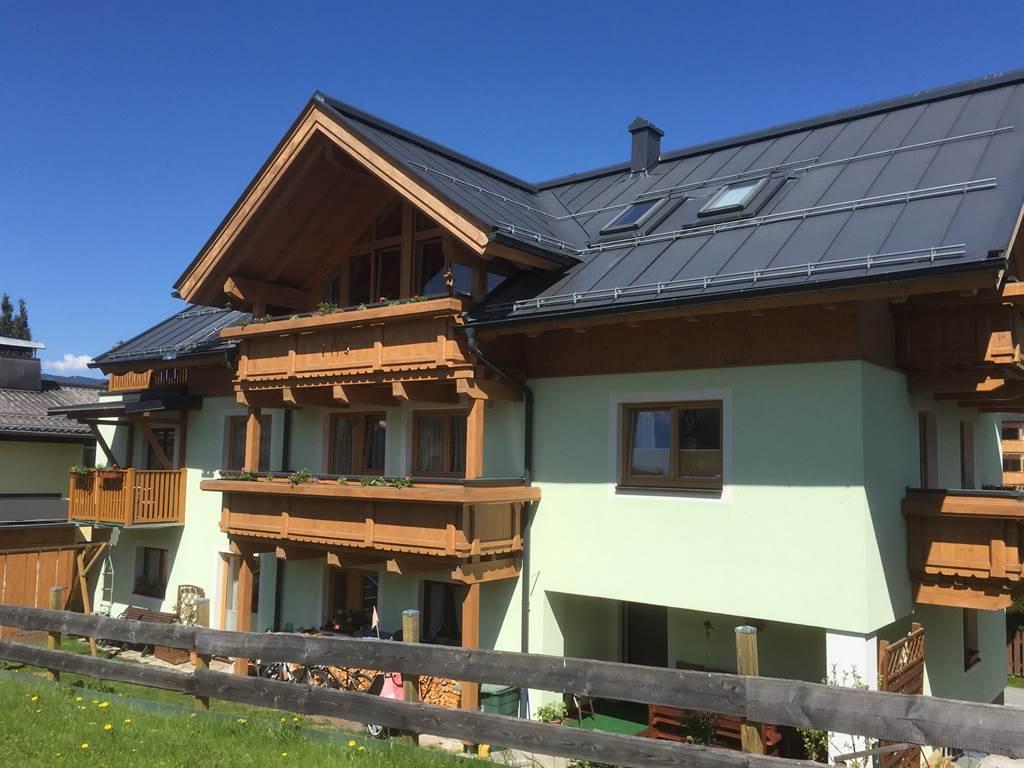 A modern house in Alpine style with wooden balustrades and a green-painted ground floor. The clear blue sky and the surrounding landscape give the scene an idyllic atmosphere.