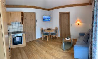 A cozy apartment with a small kitchen, a dining table, and a sofa. The walls are light and the floors are made of wood.