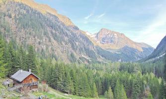 A picturesque mountain landscape with a wooden cabin and dense, green forests. In the background, majestic mountains rise into the clear sky.