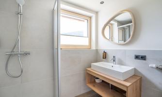 A modern bathroom with a shower, a rectangular sink, and a wooden shelf. Large windows provide plenty of daylight.