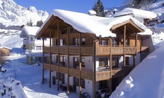 A cozy wooden house in the snow, surrounded by impressive mountains. The clear sky and winter landscape create a tranquil atmosphere.