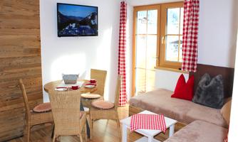 A cozy living kitchen with a rattan dining table and a couch. The windows with red checkered fabrics let in a lot of light and provide a view outside.