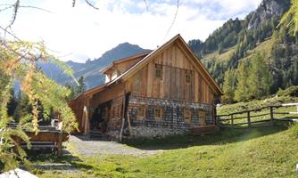 A rustic mountain cabin surrounded by green meadows and mountains. The sun shines on the wooden building and the scenery is picturesque.