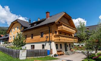 A traditional wooden house with two floors and balconies. Surrounded by green countryside and blue skies.