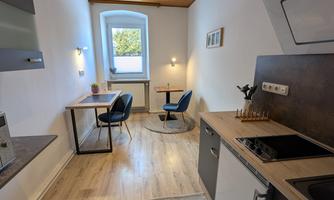 A modern kitchen with bright wooden floors and a large window. To the right are a dining table and two cozy chairs.