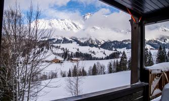 A snowy landscape with high mountains and clouds in the background. In the foreground, you can see trees and a rustic wooden building.