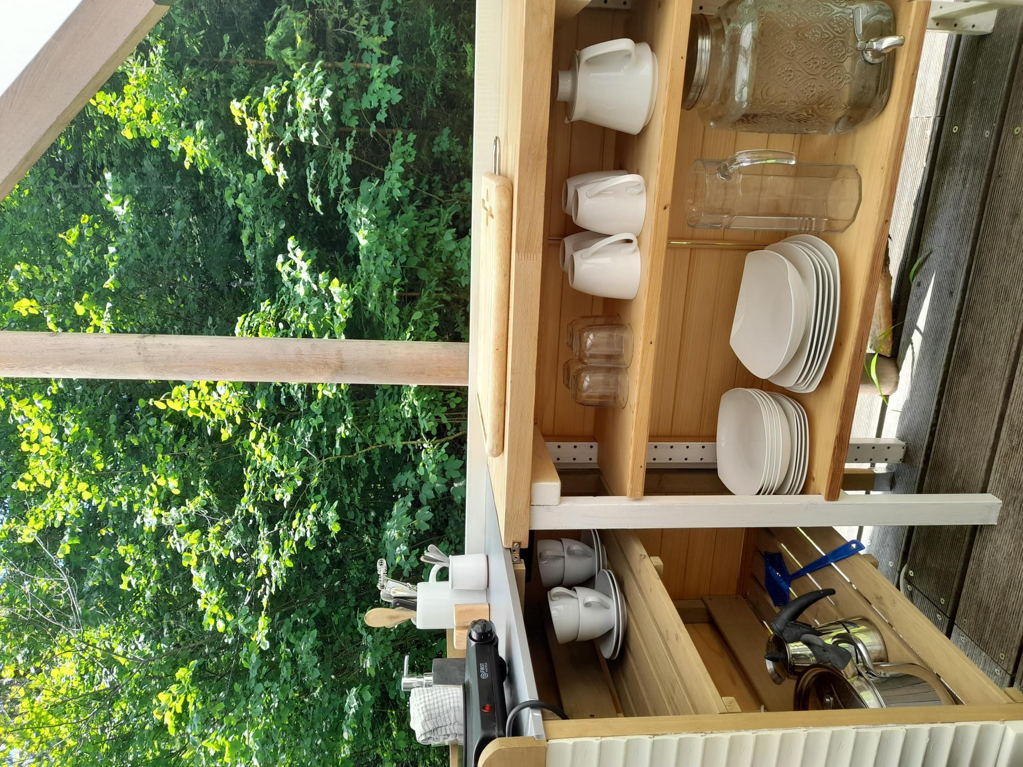 A simple outdoor kitchenette with dishes and pots. In the background, green trees can be seen.