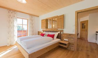 A modern bedroom with a double bed and wooden paneling. Large windows let in plenty of daylight.