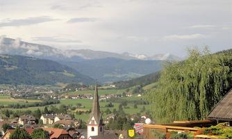 A picturesque village in a mountainous landscape. In the foreground stands a church tower, surrounded by green hills and small houses.