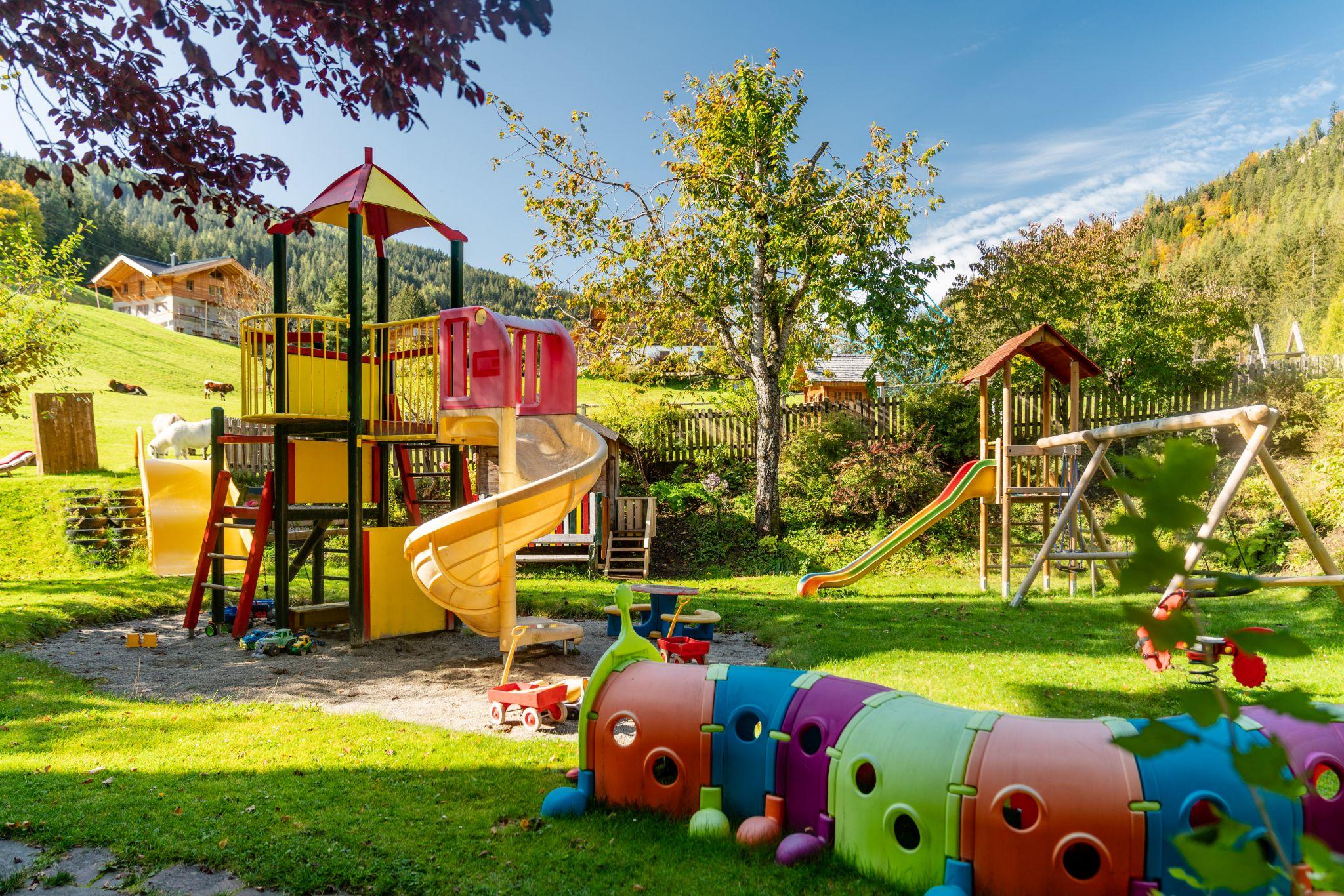 A colorful playground with slides and climbing frames surrounded by trees. In the foreground is a colorful play tunnel for children.