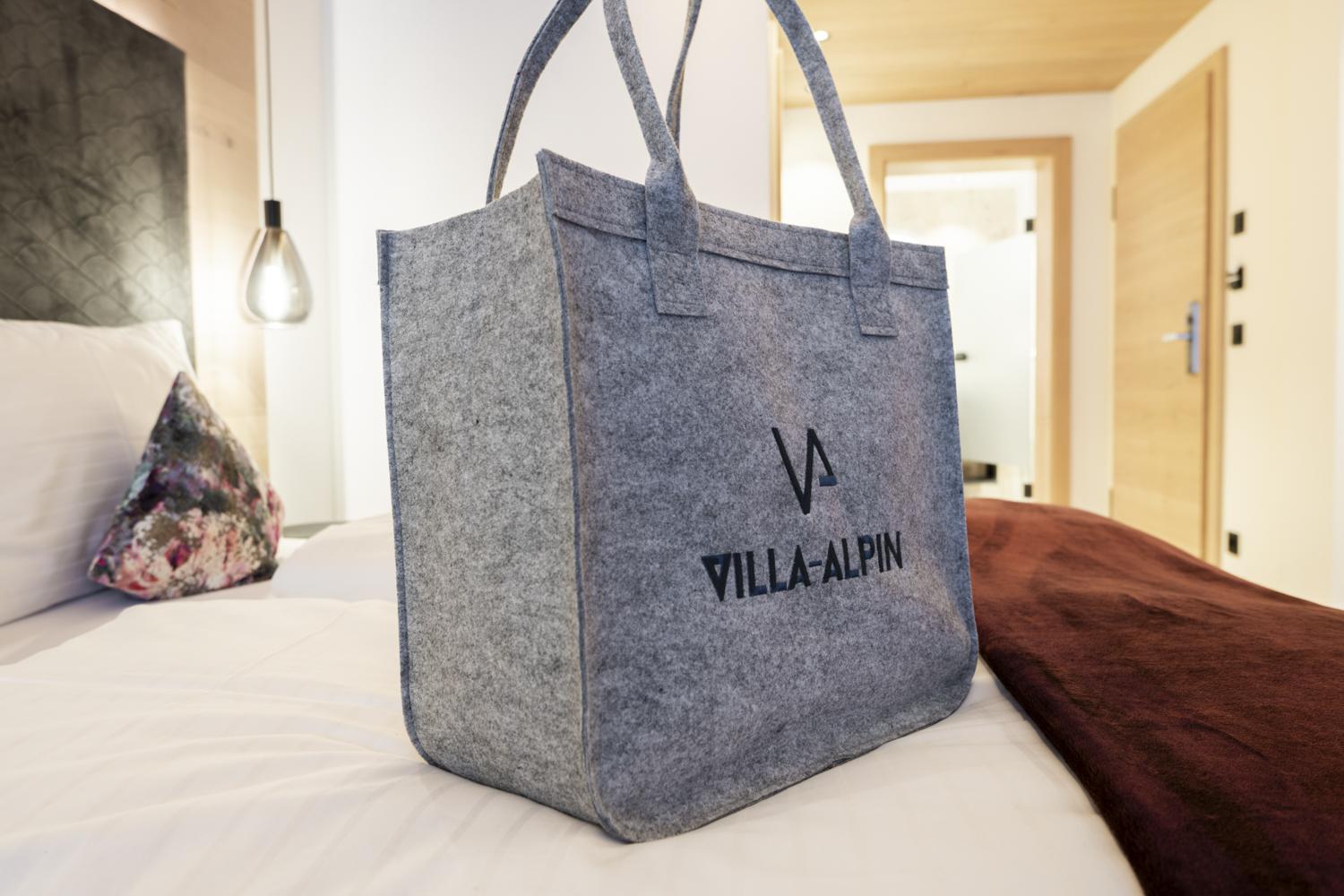 A timeless felt bag with the logo "VILLA-ALPIN". It sits on a bed in a stylish, modern room.