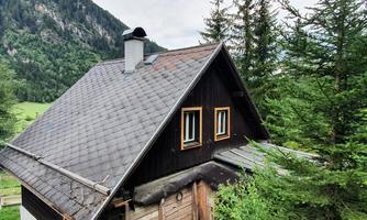 A charming wooden house surrounded by green trees and mountains. The sky is partly cloudy and the landscape appears scenic.