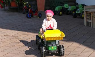 A small child is driving a yellow toy tractor over a paved floor. In the background, more toys and plants can be seen.