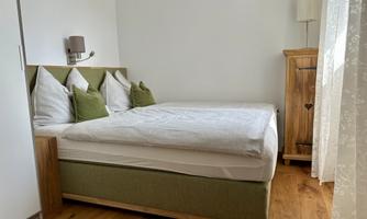 A cozy bedroom with a box spring bed and green pillows. The room features wooden flooring and a warm, inviting atmosphere.
