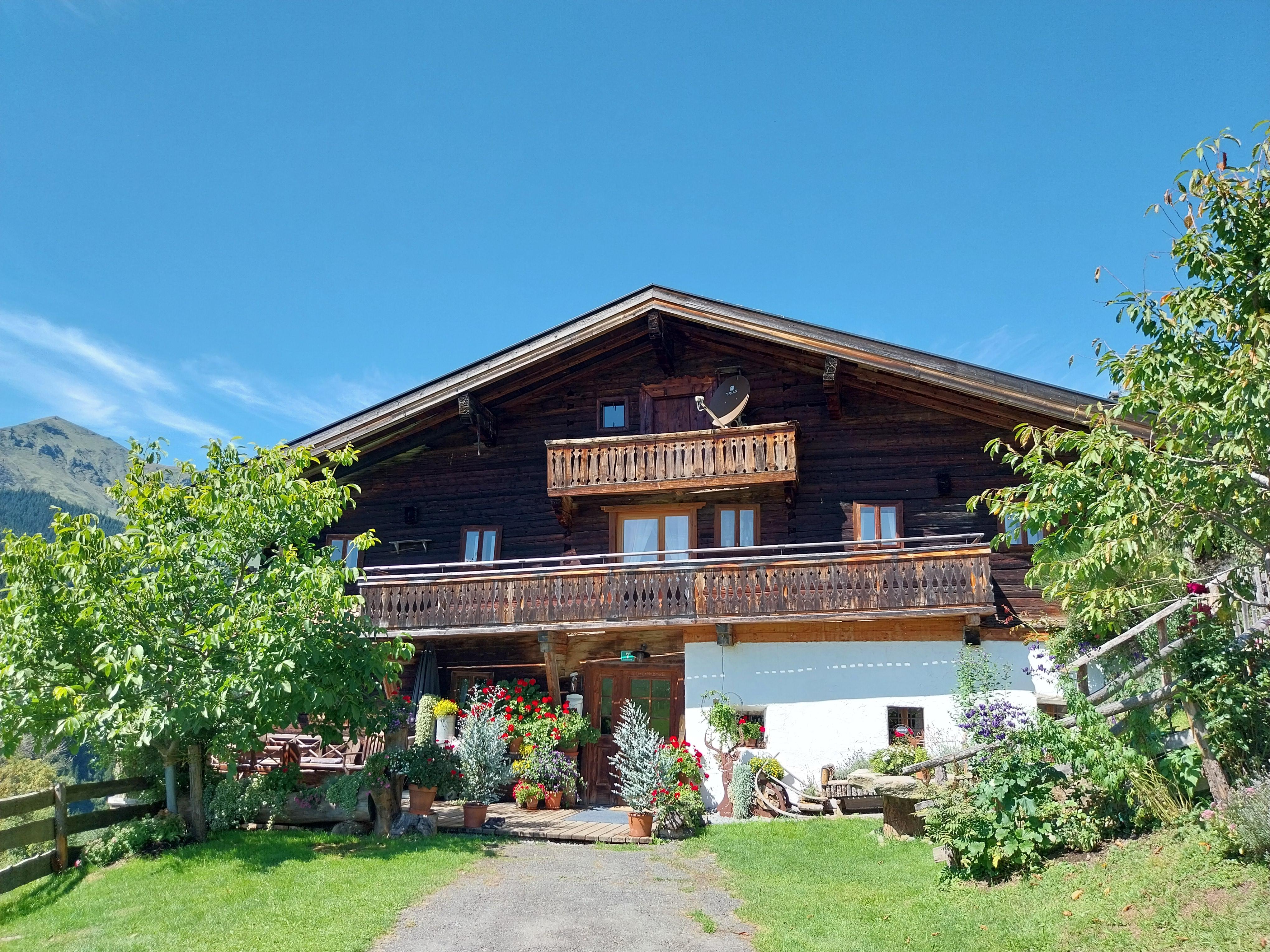 A traditional wooden house with a balcony and colorful flowers in front of the entrance. Surrounded by green trees and a clear blue sky.