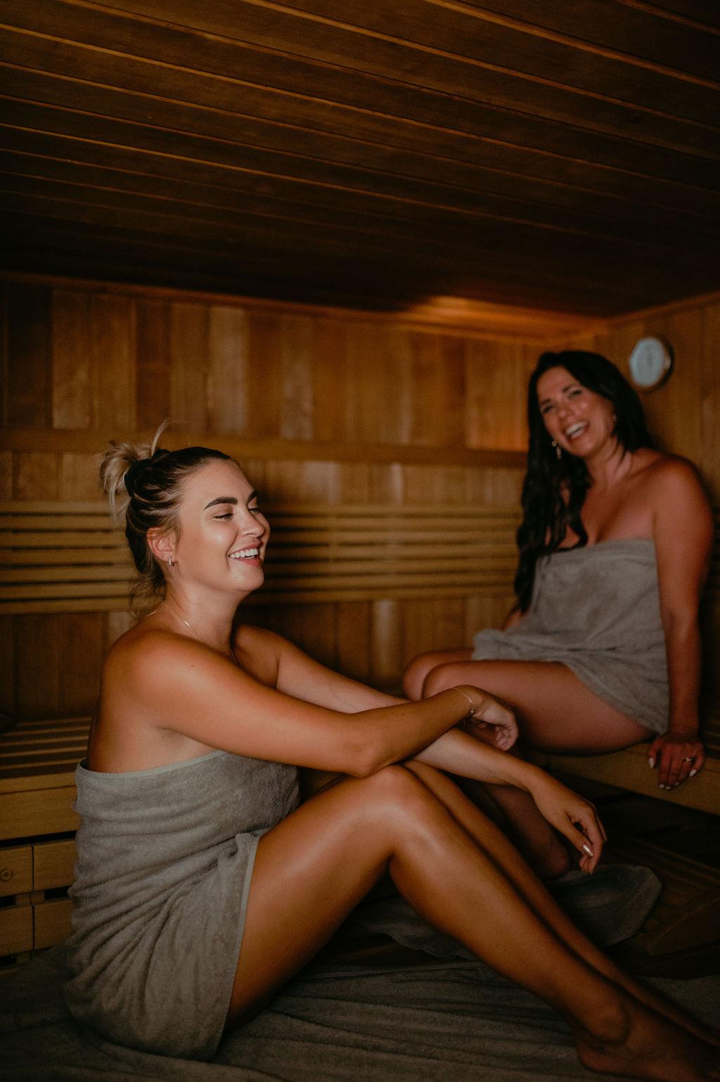 Two women are sitting relaxed in a sauna. They are wearing towels and smiling happily.