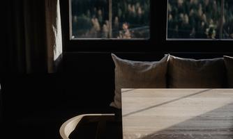 A cozy room with a wooden table and cushions on a bench. The light falls softly through the window, illuminating the surroundings.